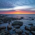 the sun is setting over the ocean with rocks in the foreground